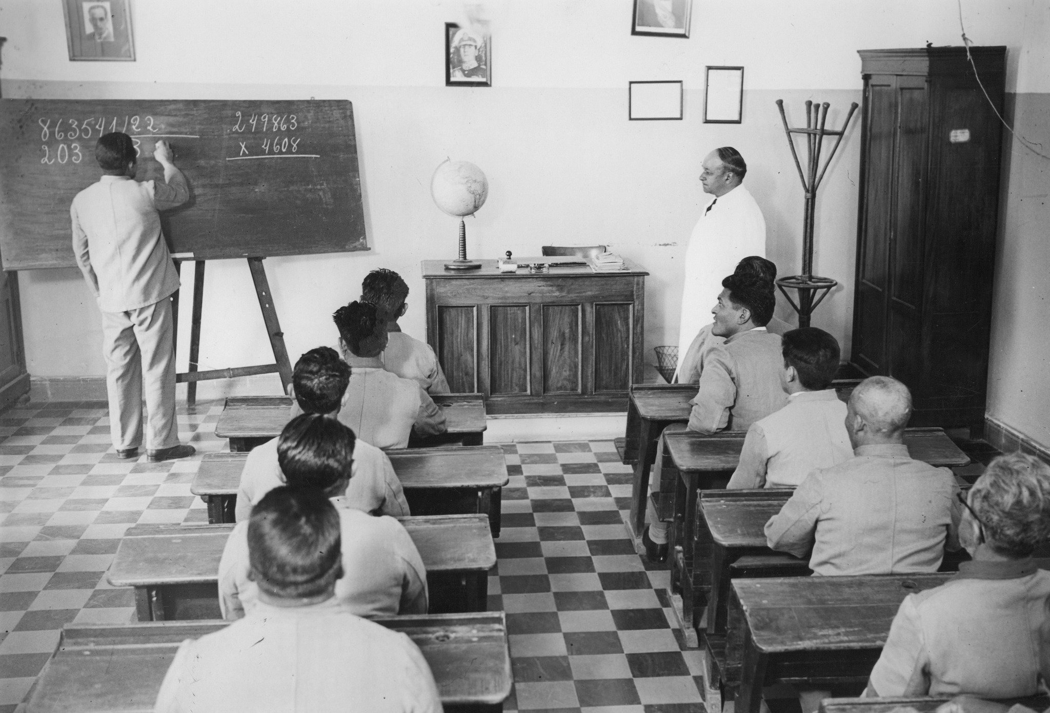 Aula de una institución penitenciaria. Los bancos alineados, el pizarrón, el globo terráqueo y la posición del instructor reúnen enseñanza y vigilancia: cada interno ocupa un lugar visible y uno de ellos expone su desempeño ante el grupo.